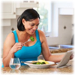 Photo of a woman enjoying a healthy lunch at her kitchen table while browsing on a tablet