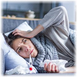 Photo of a woman with flu symptoms laying on a couch wrapped in blankets and warm clothes, holding a crumpled tissue in her hand and a thermometer in her mouth