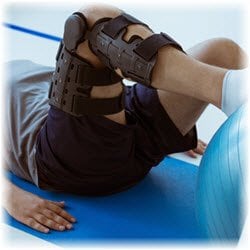 Photo of a man wearing a leg brace laying on the ground with his leg pushing against an exercise ball